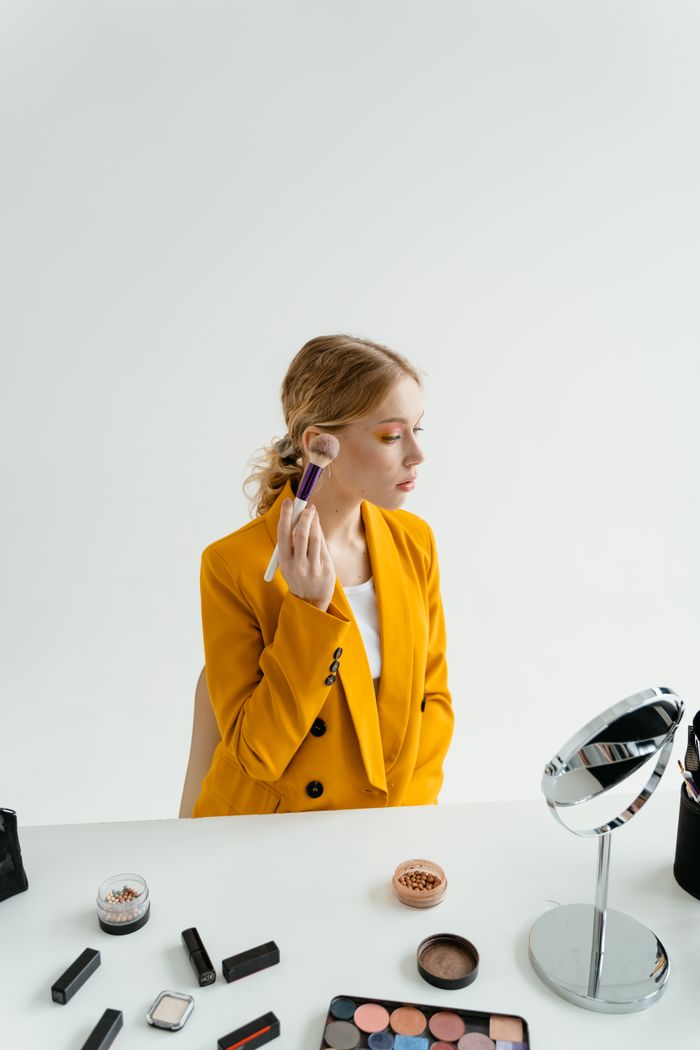 Mastering the First Impression: Your intriguing post title goes here Woman in yellow blazer applying makeup using a brush at a vanity in a studio setting.